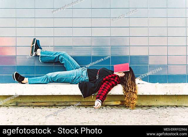 Teenage girl lying on a wall outdoors covering face with book