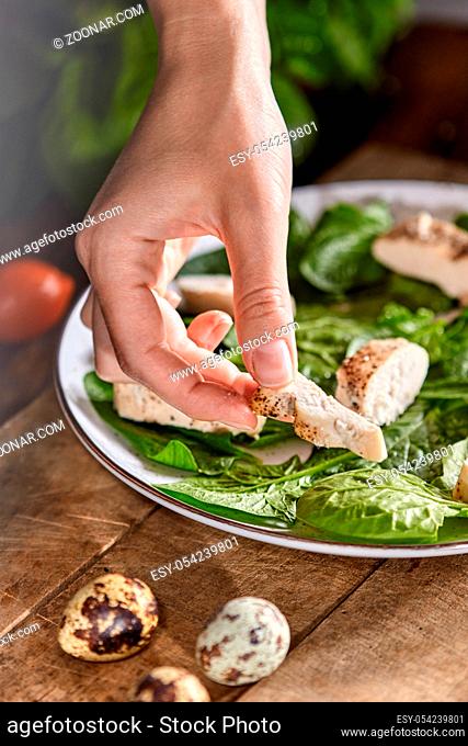 A plate with spinach on a wooden table. Woman's hand puts a piece of meat in the plate. Step by step cooking healthy dietary salad