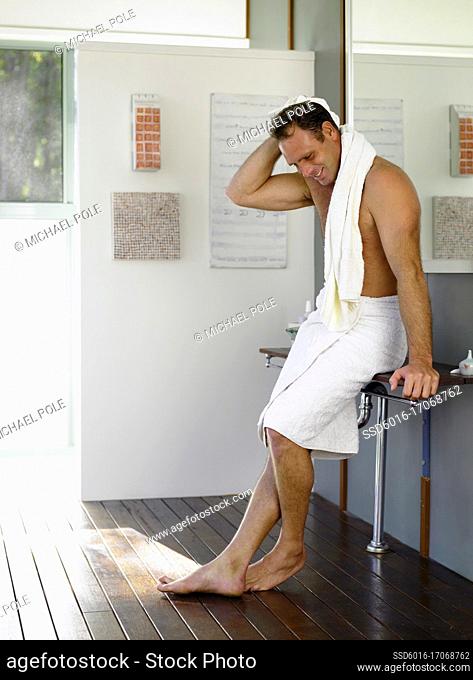 Man with towel wrapped agound his waist and towell drying his hair, leaning on bench in bathroom