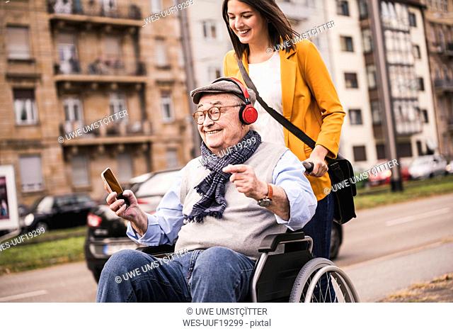 Laughing young woman pushing happy senior man with headphones and smartphone in wheelchair