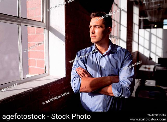 Thoughtful caucasian businessman standing in sunny office with arms crossed looking out of window
