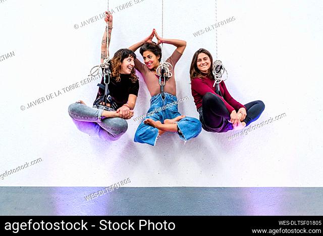 Smiling female acrobats hanging on rope with cross-legged in front of wall