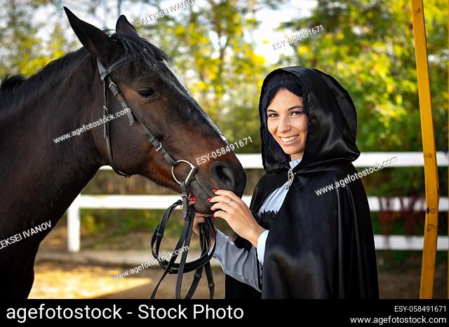 Happy girl in medieval clothes and a black cloak stands with a horse horse on the background of trees and a fence