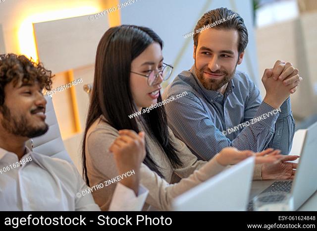 Focused young company employee pointing at something on the laptop to her pleased male colleagues