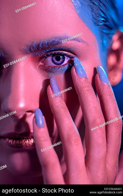 Young stylish girl with bright makeup with a hand near the face. In the neon shade the face of the model. near the face is a hand with blue nails