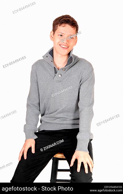 A smiling young handsome teenage boy sitting in a gray sweater on a chair, isolated for white background