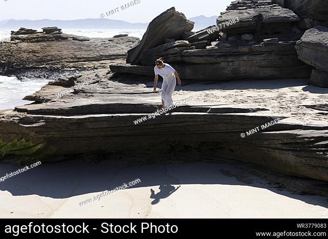 Teenage girl preparing to jump from smooth flat layered rocks above a sandy beach