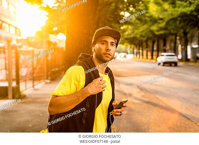 Portrait of young man listening music with earphones and smartphone waiting at roadside