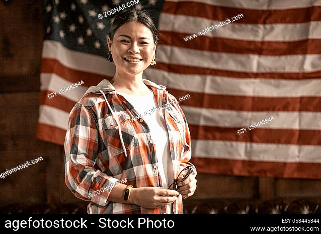 Beautiful Asian Woman Feels Free Rejoicing In Independence. The Brunette Middle-Aged Holds Her Glasses And Cheerful Laughs At The Striped Flag Of America...