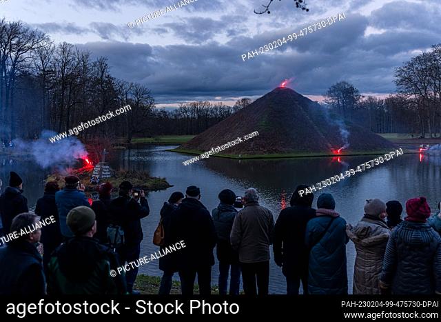 04 February 2023, Brandenburg, Cottbus: The lake pyramid in Pückler Park Cottbus-Branitz and an island with a memorial stone are illuminated with Bengal fire on...