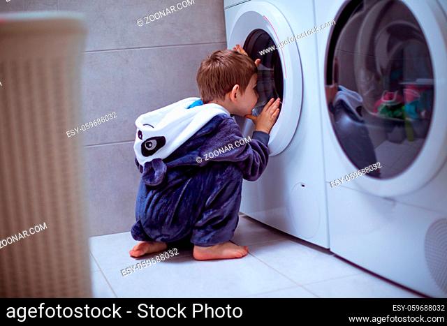 Back view of a baby is sitting on the floor and looking into the washing machine. The kid in soft pajamas carefully watches washing