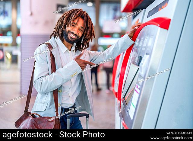 Happy businessman with luggage pointing at ticket machine in station