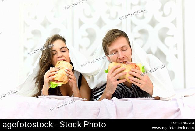 Young couple eating delicious hamburgers while lying in bed covered. People resting and relaxing after hard working day or week