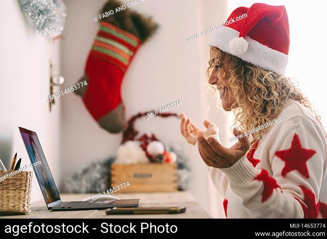 Happy woman using laptop at home to video call a friend during christmas holiday. Female people side portrait with santa claus hat and xmas decorations in...
