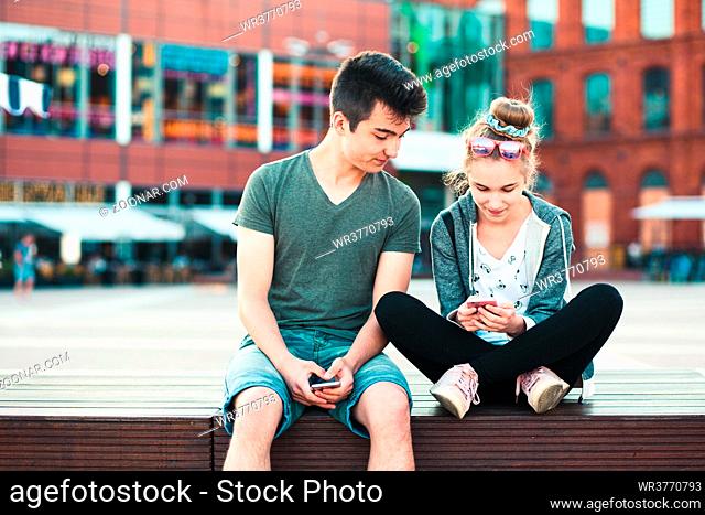 Couple of friends, teenage girl and boy, having fun together with smartphones, sitting in center of town, spending time together