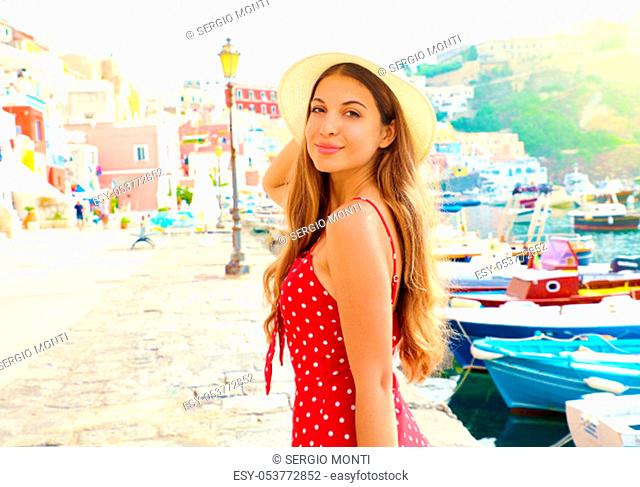 Beautiful young woman in Procida, Italy. Fashion girl with red dress and hat walks along the harbor of Procida Island in Italy