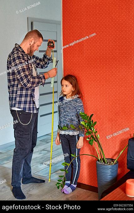 Father measures daughter's height against wall at home, family lifestyle. Copy space