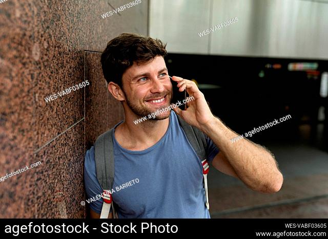 Happy man talking through mobile phone while looking away and leaning on brown tile wall at subway station