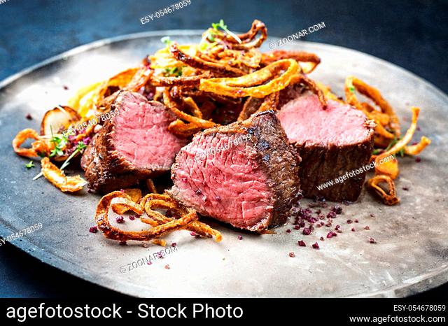 Traditional dry aged sliced beef tenderloin with fried onion rings and potatoes served as closeup on a modern design plate