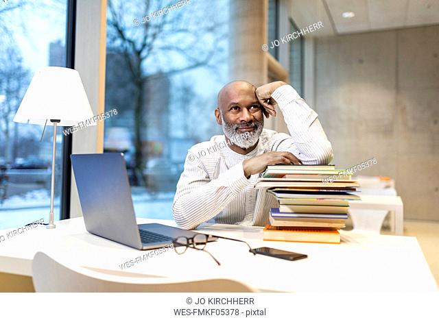Portrait of smiling mature man sitting at desk with laptop and stack of books