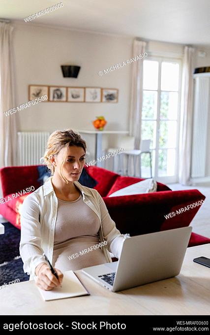 Female freelancer writing in book while using laptop at desk in living room