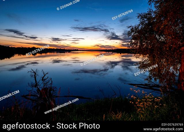 Summer evening twilight view on picturesque plain lake with forest and blossoming meadows on shores and sunset clouds reflections in water surface