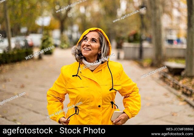 Thoughtful senior woman wearing yellow raincoat standing at footpath