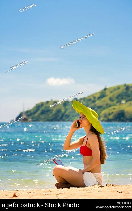 Fashionable young woman talking on mobile phone while using a tablet on the beach in a sunny day of summer in Flores Island, Indonesia