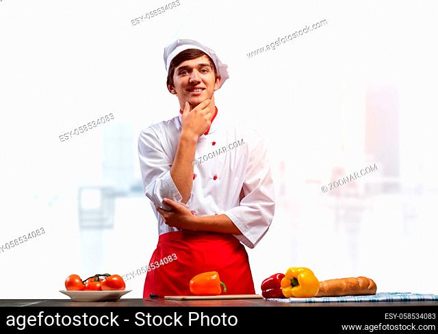 Young smiling chef standing near cooking table with fresh vegetables. Pleased chef in white hat and red apron in light kitchen interior