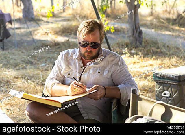 Bearded man wearing sunglasses sitting on chair in a safari camp, writing a journal
