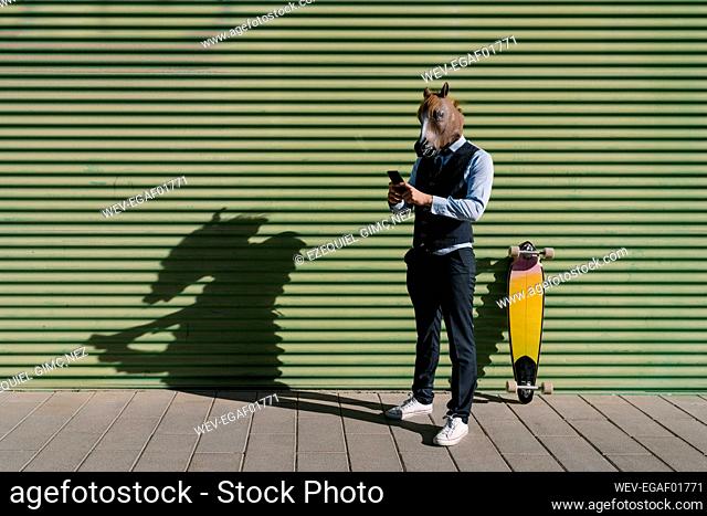 Male entrepreneur in horse mask using mobile phone while standing against wall