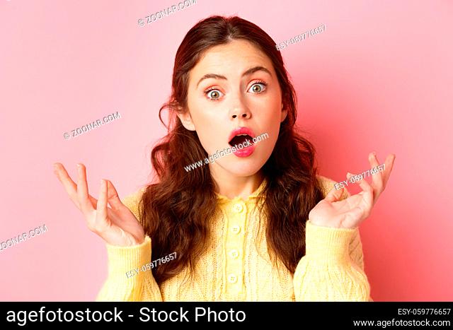 Close up of surprised and confused young woman open mouth and raise hands, stare at camera puzzled, cant understand something, standing against pink background