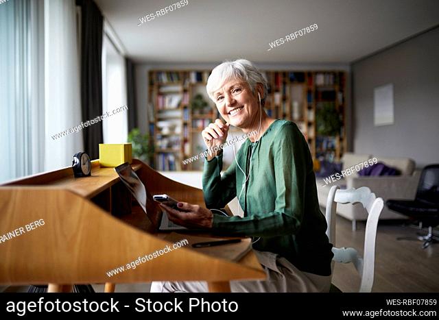 Smiling active senior woman listening music while sitting on chair at home