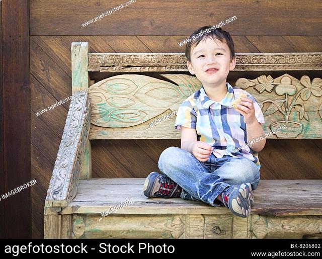 Cute relaxed mixed-race boy sitting on bench eating his sandwich