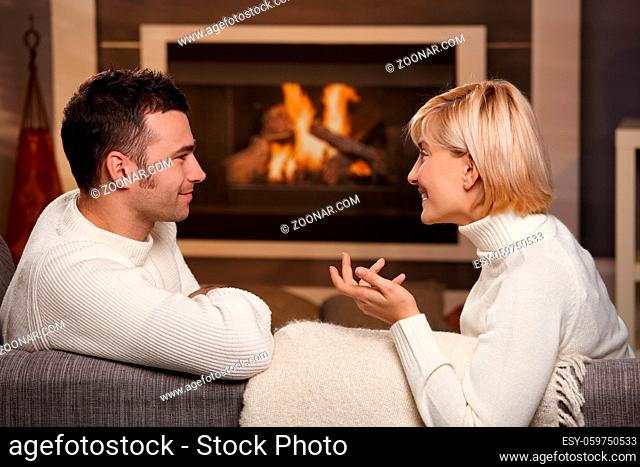Young romantic couple sitting on sofa in front of fireplace at home, looking at each other, talking