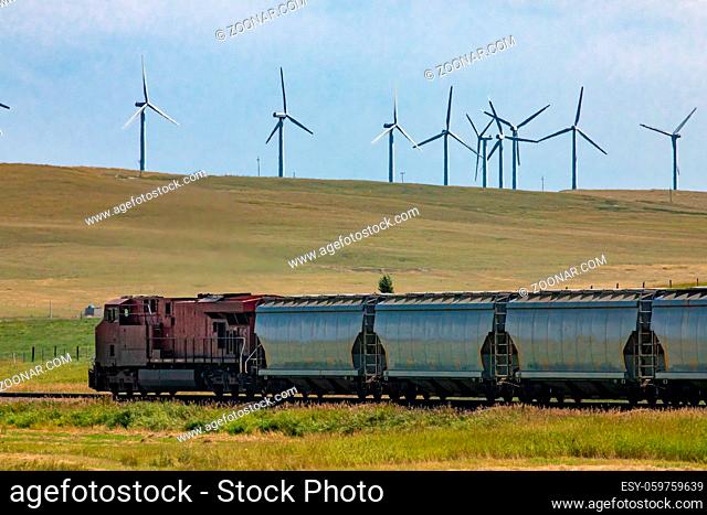 Row of metallic wagons of a Canadian freight train running between green fields in the countryside. Wind turbines in the background. Red locomotive
