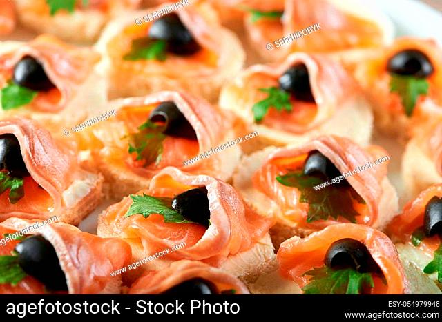 Little canapees with smoked salmon rolls and black olives on the white bread, covered with butter and decorated with fresh green parsley leaves