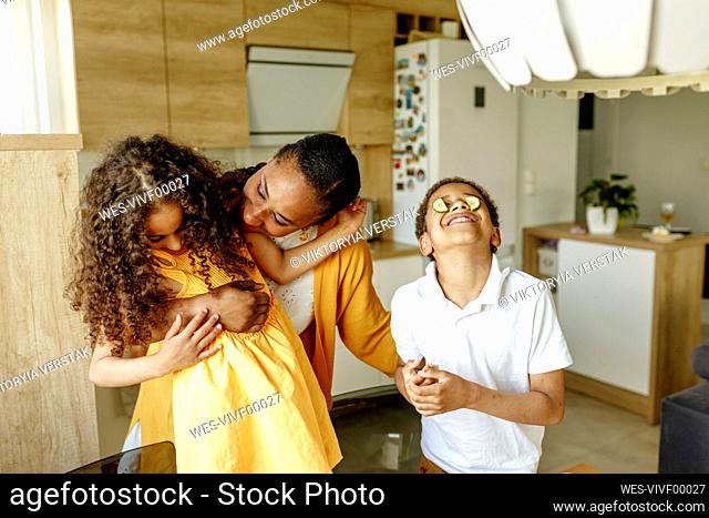 Happy mother embracing daughter by son covering eyes with cucumber in kitchen