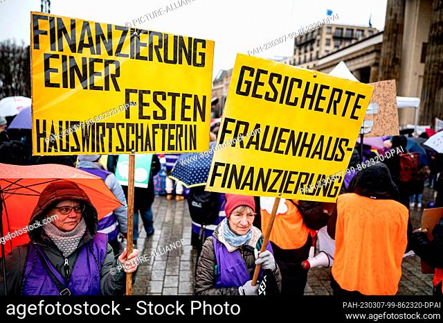 07 March 2023, Berlin: A participant stands with signs reading ""Funding for a permanent housekeeper"" and ""Secured women's shelter funding"" at a protest...