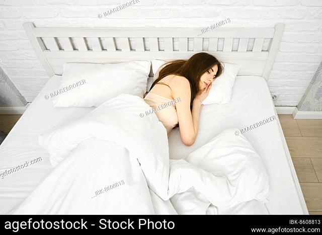 Young cute woman peacefully sleeping on comfortable bed with fresh and clean white bedding, female having sweet dreams while resting on soft pillow in bedroom