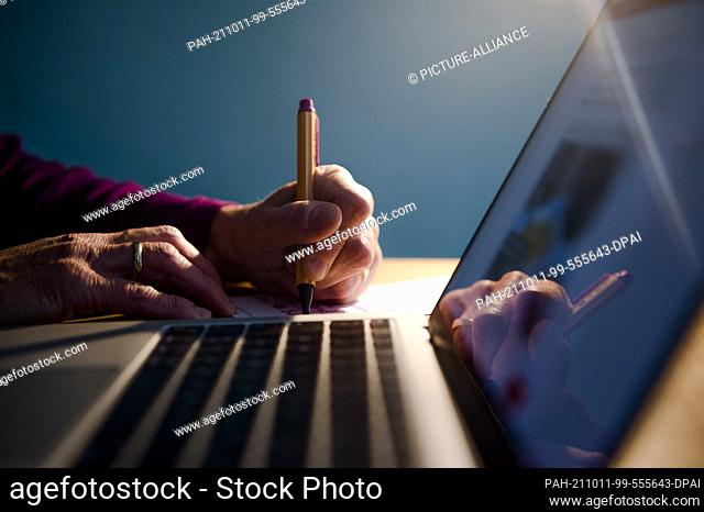 PRODUCTION - 01 October 2021, Bavaria, Munich: A woman is sitting at a desk at home. She writes with a pen on a sheet of paper next to a laptop