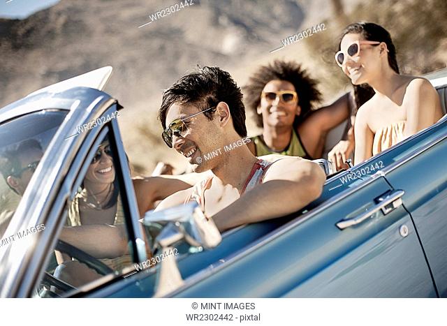 A group of friends in a pale blue convertible on the open road, driving across a dry flat plain surrounded by mountains