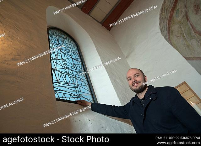09 October 2021, Saxony-Anhalt, Altjeßnitz: Artist Bastian Muhr stands next to one of the modern church windows he designed in the small Romanesque church in...