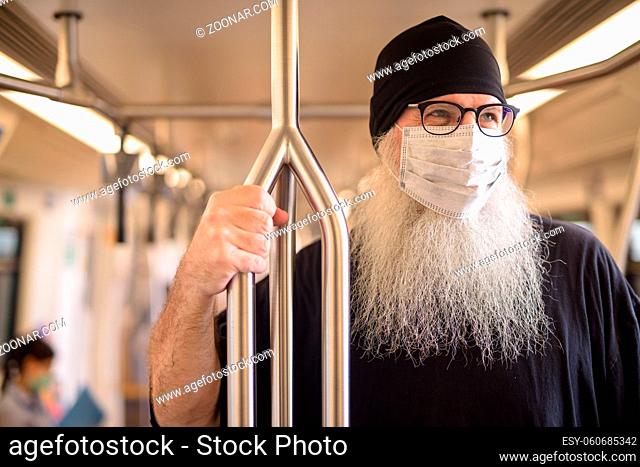 Portrait of mature bearded hipster man as tourist with mask for protection from corona virus outbreak inside the train