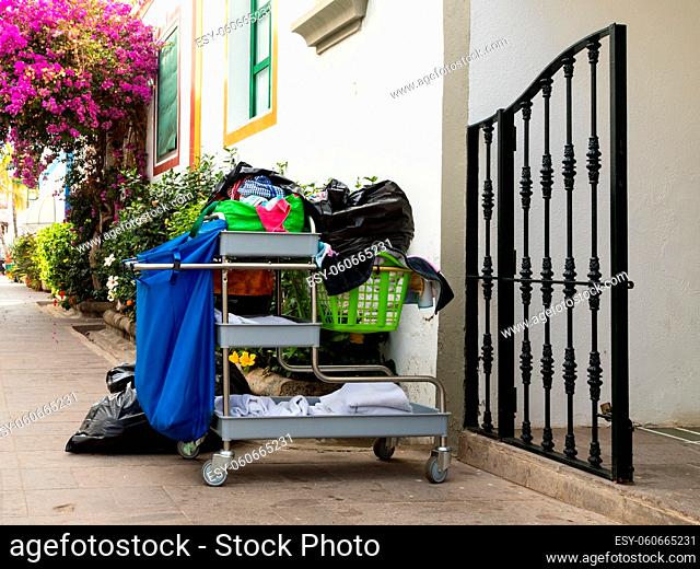 Cleaning trolley with tools and towels, standing outside a gate in the street, with colorful flowers in the background. Puerto de Mogan, Gran Canaria in Spain