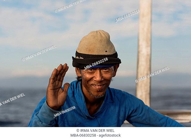 Friendly, balinese fisherman sits in his Jukung (balinese outrigger canoe) and smiles friendly into the camera, lifts his right hand