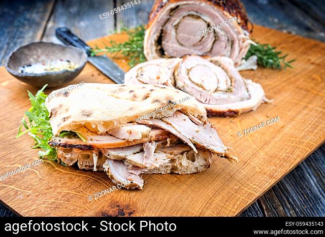 Traditional Italian Porchetta arrotolata pork meat sliced and as piece with pita bread and rocket salad offered as close-up on a modern design wooden board