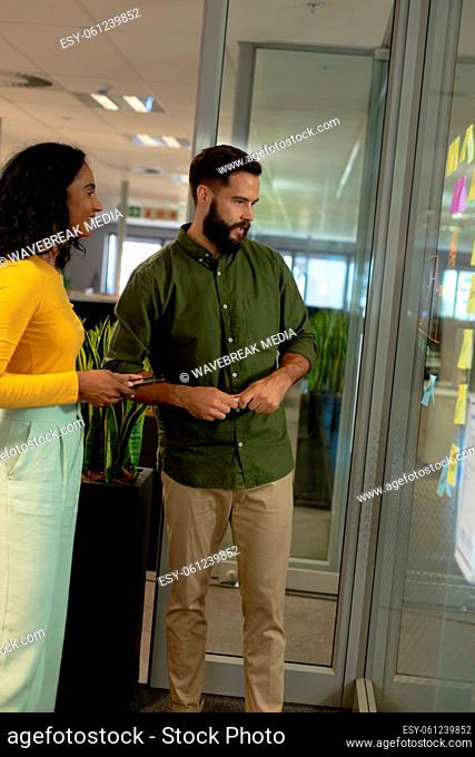 Biracial business colleagues discussing while looking at strategy on glass wall in workplace