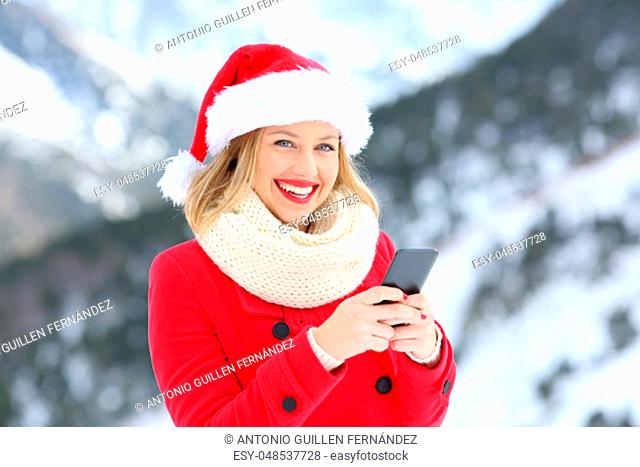 Portrait of a happy woman on christmas holidays using phone looking at you with a snowy mountain in the background