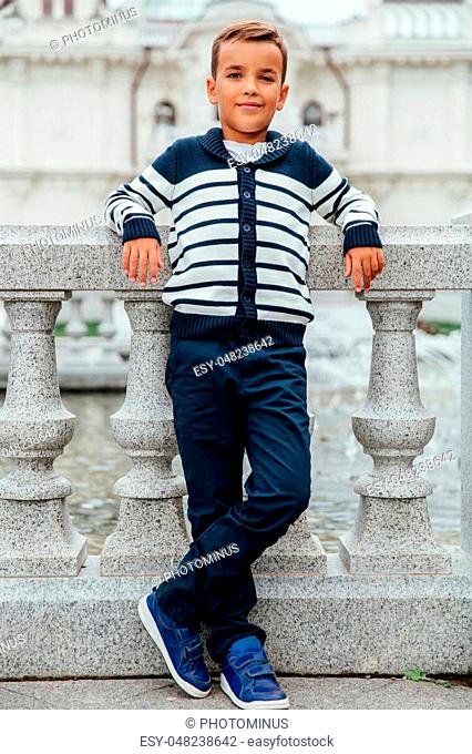 Stylish rich little boy in fashionable clothes . Leaned on Granite-stone handrails. stylish, handsome posing outside in the city park
